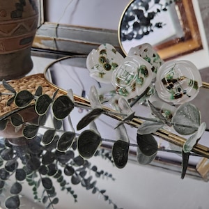 May include: A collection of translucent white resin buttons featuring embedded dried green and brown botanical elements. The buttons are arranged on a gold-rimmed hexagonal mirror, accompanied by a sprig of artificial eucalyptus with grey-green leaves.