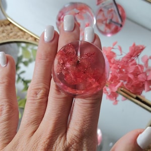 May include: A circular resin ring featuring a clear, transparent finish with embedded dried red hydrangea petals. The ring has a smooth, rounded surface and a polished appearance, showcasing the natural texture of the preserved floral elements.