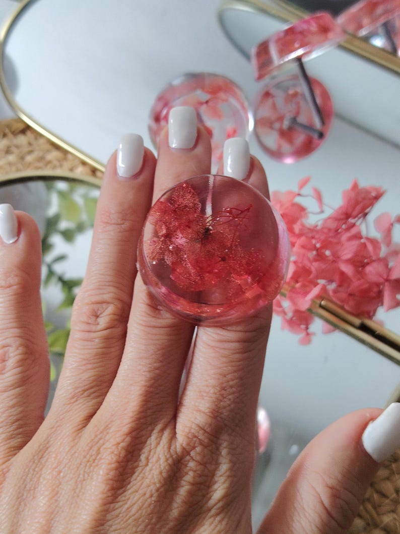 May include: A circular resin ring featuring a clear, transparent finish with embedded dried red hydrangea petals. The ring has a smooth, rounded surface and a polished appearance, showcasing the natural texture of the preserved floral elements.