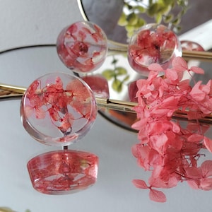 May include: Three clear, circular resin cabochons containing dried pink hydrangea petals. The resin pieces are smooth and transparent, showcasing the delicate structure of the preserved flowers inside. A cluster of loose pink hydrangea petals sits nearby.