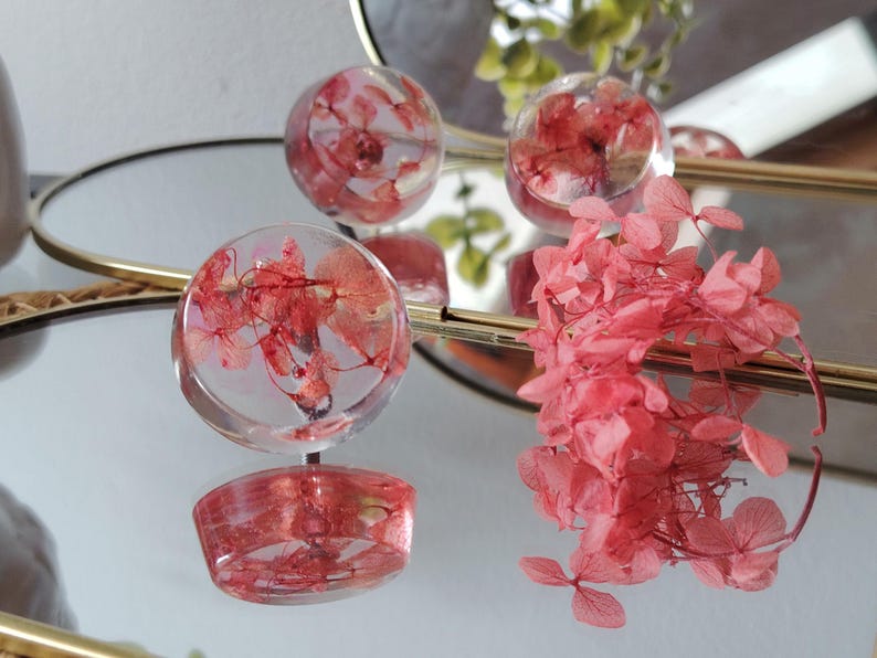 May include: Three clear, circular resin cabochons containing dried pink hydrangea petals. The resin pieces are smooth and transparent, showcasing the delicate structure of the preserved flowers inside. A cluster of loose pink hydrangea petals sits nearby.