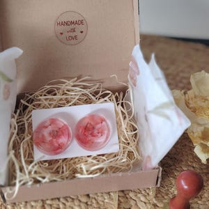 May include: A pair of round stud earrings featuring embedded pink and white flower petals within clear resin, presented on a white card inside a small brown cardboard box with a sticker that reads HANDMADE with LOVE.