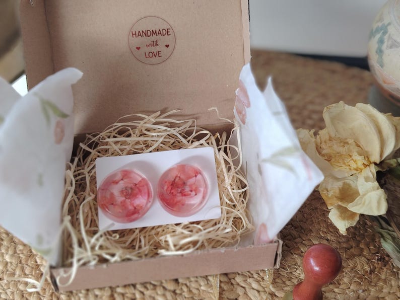 May include: A pair of round stud earrings featuring embedded pink and white flower petals within clear resin, presented on a white card inside a small brown cardboard box with a sticker that reads HANDMADE with LOVE.