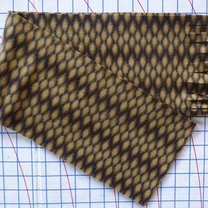 May include: A brown and tan fabric strip with a diamond pattern and frayed edges. The fabric is measured on a white grid with blue and red lines.