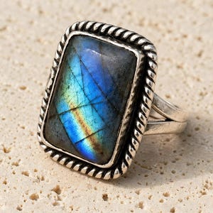 May include: A rectangular labradorite gemstone ring featuring a polished, iridescent blue and green stone set in a silver-toned metal frame with a twisted rope-style border and a split shank band design.