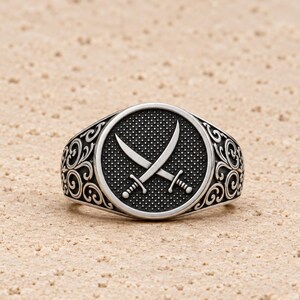 May include: A silver-toned ring featuring a circular face with a stippled background and two crossed swords. The band is decorated with intricate scrollwork patterns on both sides. The text OVR CREATIONS is visible in the bottom left corner.