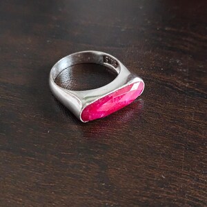 May include: A silver ring featuring a horizontal, elongated, faceted hot pink gemstone set into the band. The interior of the silver band is clearly stamped with the text 925, indicating sterling silver composition for this piece of jewelry.