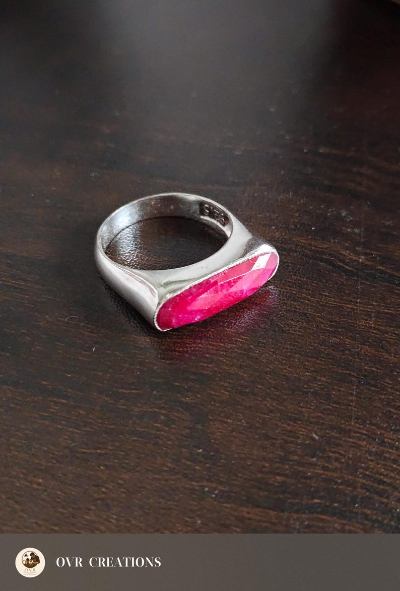 May include: A silver ring featuring a horizontal, elongated, faceted hot pink gemstone set into the band. The interior of the silver band is clearly stamped with the text 925, indicating sterling silver composition for this piece of jewelry.