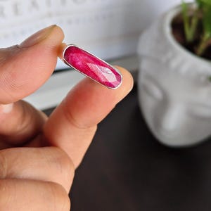 May include: A ring featuring a long, rectangular, faceted deep pink ruby gemstone set in a smooth, polished silver metal bezel. The stone has a vibrant, saturated color with visible natural inclusions and a flat, elongated horizontal orientation.