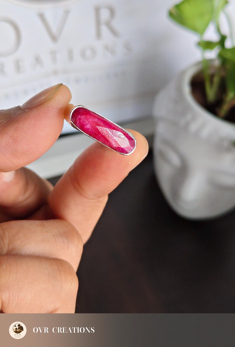 May include: A ring featuring a long, rectangular, faceted deep pink ruby gemstone set in a smooth, polished silver metal bezel. The stone has a vibrant, saturated color with visible natural inclusions and a flat, elongated horizontal orientation.