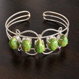 Peut inclure: Un bracelet manchette en argent orné de cinq pierres vertes en forme de goutte avec des motifs de matrice brune. La bande métallique présente une finition texturée et un design en filigrane délicat maintenant chaque pierre en serti griffe.