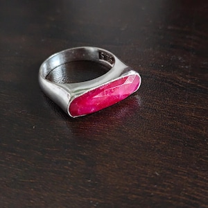 May include: A silver-toned ring featuring a horizontal, elongated, faceted hot pink gemstone set into the band. The interior of the ring band is clearly stamped with the text 925, indicating the metal composition of the jewelry piece.