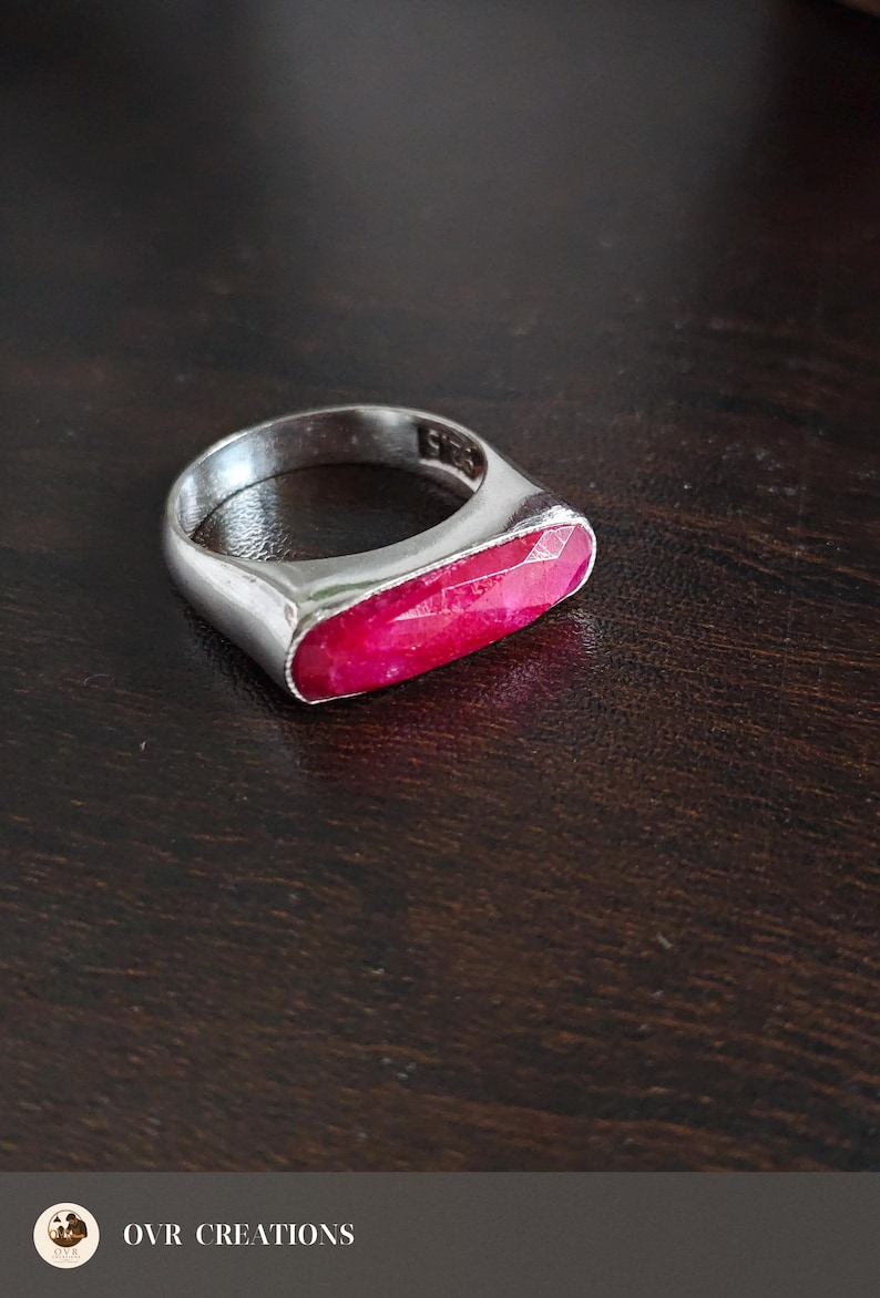 May include: A silver-toned ring featuring a horizontal, elongated, faceted hot pink gemstone set into the band. The interior of the ring band is clearly stamped with the text 925, indicating the metal composition of the jewelry piece.