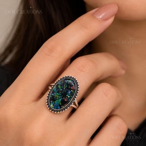 May include: An oval-shaped ring featuring a dark black inlay filled with iridescent, multi-colored flecks that shimmer in shades of green, blue, and red. The center stone is framed by a textured, rope-style silver border on a thin silver band.
