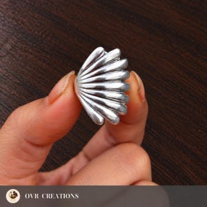 Può includere: Un anello color argento a forma di conchiglia a ventaglio con evidenti creste verticali e una finitura metallica lucida. Il design presenta un bordo curvo semicircolare con segmenti distinti che si irradiano dalla base.