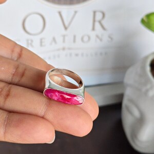 May include: A silver ring featuring a long, rectangular, faceted hot pink gemstone set horizontally. The band is smooth and polished silver, showcasing a clean, metallic finish that contrasts with the vibrant, textured surface of the stone.