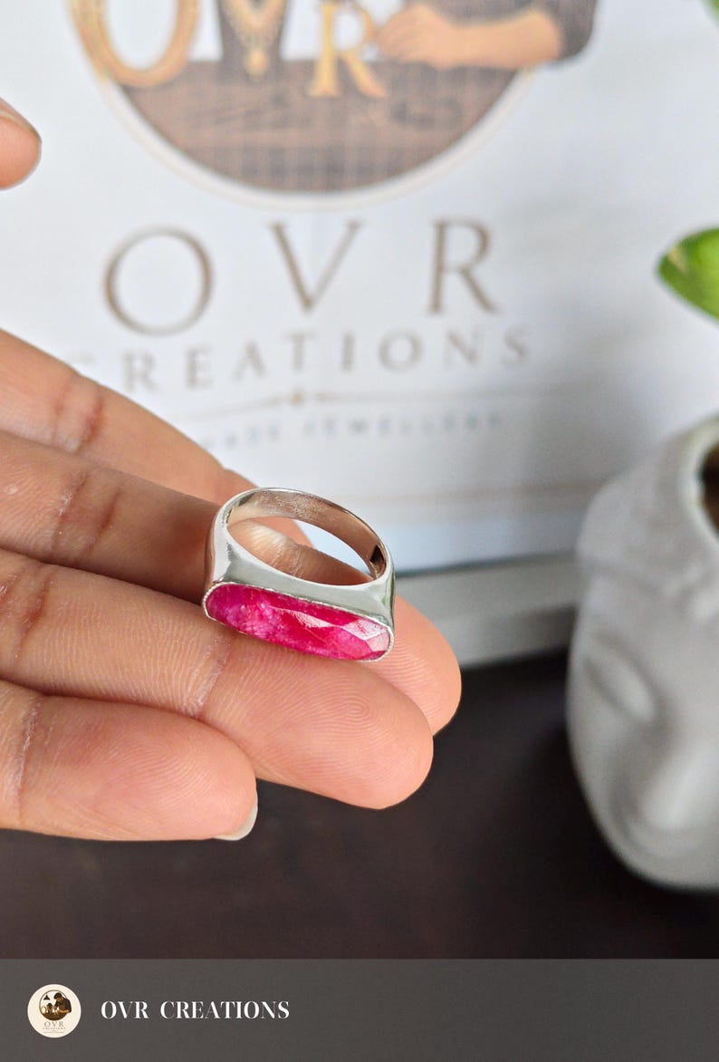 May include: A silver ring featuring a long, rectangular, faceted hot pink gemstone set horizontally. The band is smooth and polished silver, showcasing a clean, metallic finish that contrasts with the vibrant, textured surface of the stone.
