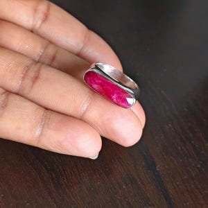May include: A silver ring featuring a long, rectangular, faceted deep pink gemstone set horizontally. The stone has a vibrant, saturated color with visible natural inclusions, and the metal band has a smooth, polished finish with a simple design.