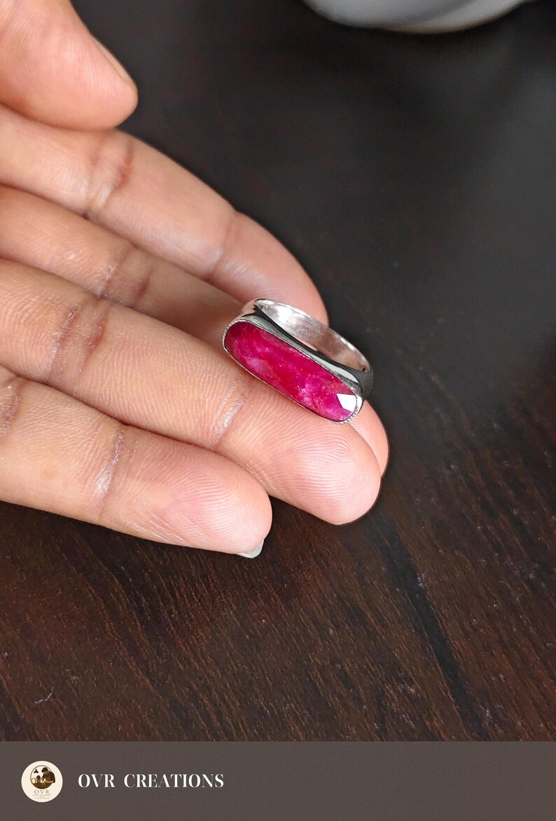May include: A silver ring featuring a long, rectangular, faceted deep pink gemstone set horizontally. The stone has a vibrant, saturated color with visible natural inclusions, and the metal band has a smooth, polished finish with a simple design.