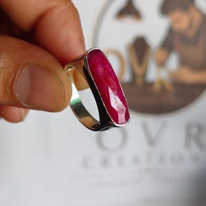 May include: A silver ring featuring a long, rectangular, faceted deep red ruby gemstone set vertically in a smooth metal bezel. The ring band is polished silver, showcasing a clean, simple design that highlights the vibrant color of the stone.