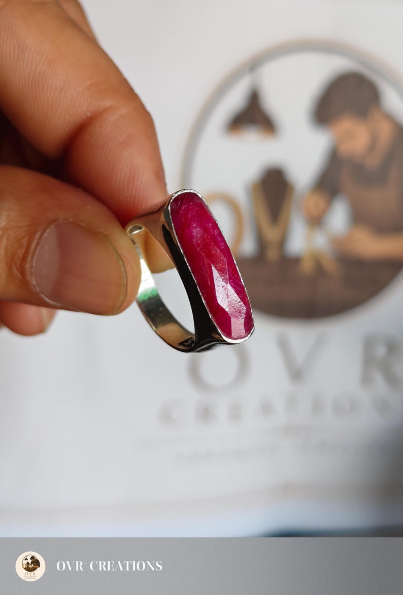 May include: A silver ring featuring a long, rectangular, faceted deep red ruby gemstone set vertically in a smooth metal bezel. The ring band is polished silver, showcasing a clean, simple design that highlights the vibrant color of the stone.