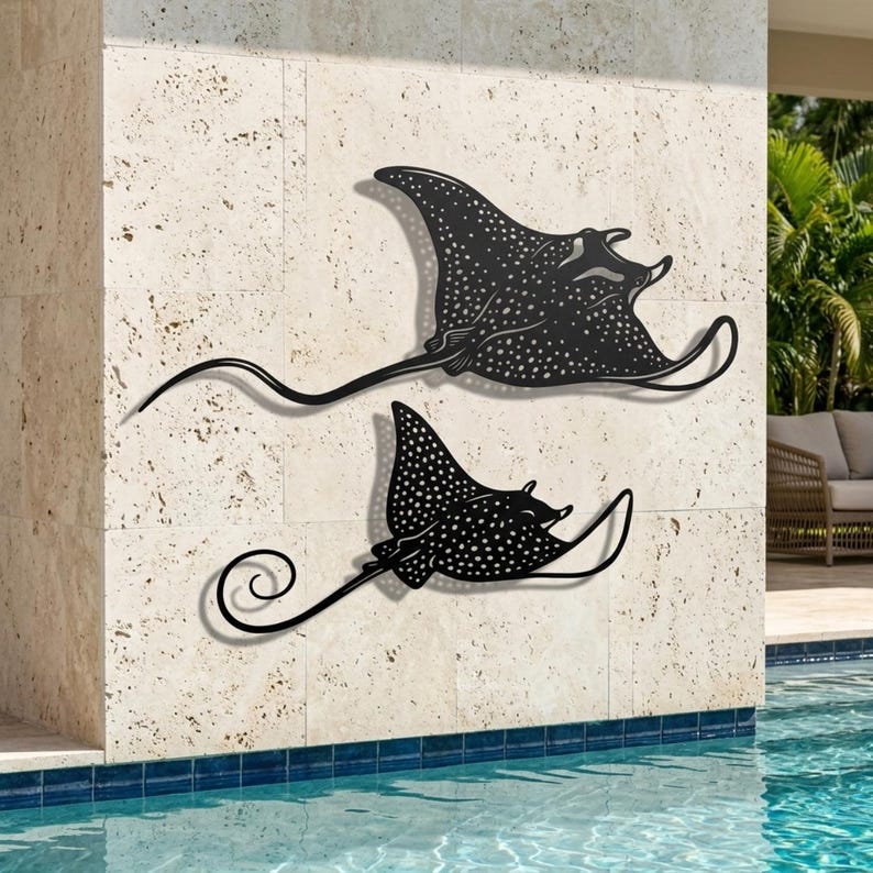May include: Two black metal wall art pieces shaped like spotted eagle rays. Each ray features a flat silhouette with a detailed white dotted pattern across the body and a long, thin, curved tail. The metal cutouts are mounted on a stone wall.