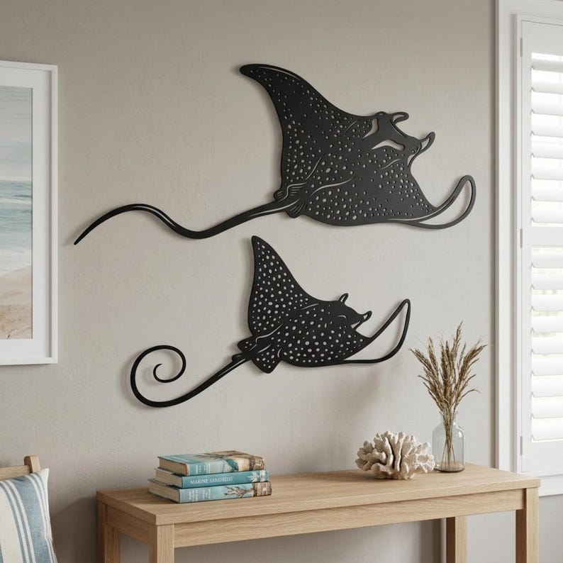 May include: Two black metal wall art pieces shaped like spotted eagle rays. Each ray features a flat silhouette with intricate cut-out patterns representing spots across the body, long flowing fins, and a slender, curved tail extending outward.