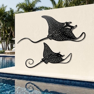 May include: Two black metal wall art pieces shaped like spotted eagle rays. Each ray features a flat silhouette with a long, slender tail and a distinct pattern of white dots across its body. The larger ray is positioned above the smaller one.