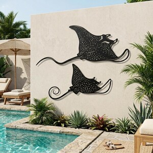 May include: Two black metal wall art pieces shaped like spotted eagle rays, featuring intricate cut-out patterns and long, curved tails. The larger ray is positioned above the smaller ray, both displaying a flat, silhouette-style design.