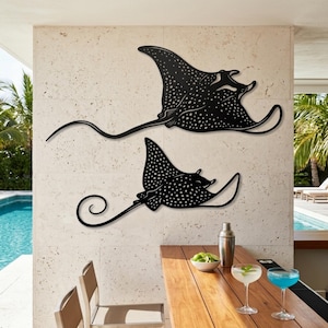 May include: Two black metal wall art pieces shaped like spotted eagle rays. Each ray features a flat silhouette with intricate cut-out white spots across the body, wide fins, and a long, slender tail. The larger ray is positioned above the smaller one.
