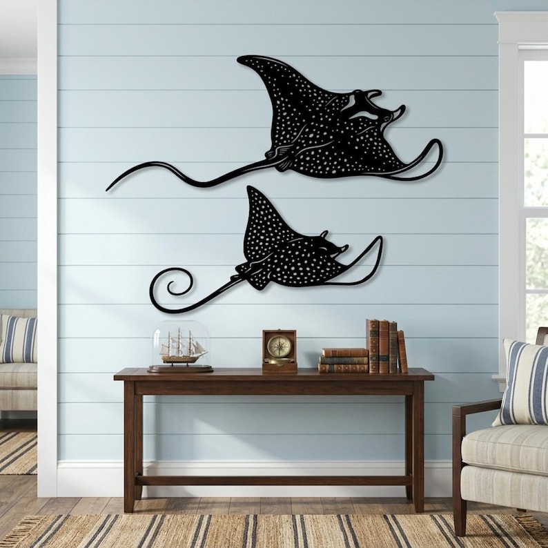 May include: Two black metal wall art pieces shaped like spotted eagle rays. Each ray features a flat silhouette with a long, thin tail and a distinct pattern of white dots across its body. The larger ray is positioned above the smaller one.