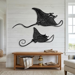 May include: Two black metal wall art pieces shaped like spotted eagle rays. The larger ray is positioned above the smaller one, both featuring intricate white dot patterns across their bodies, wide wings, and long, slender, curved tails.