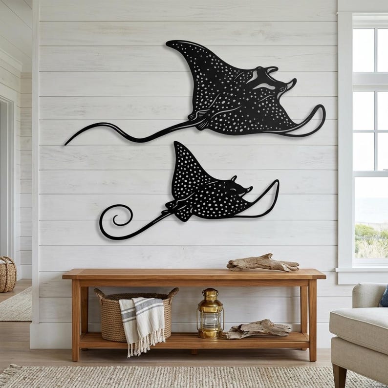 May include: Two black metal wall art pieces shaped like spotted eagle rays. The larger ray is positioned above the smaller one, both featuring intricate white dot patterns across their bodies, wide wings, and long, slender, curved tails.