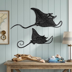 May include: Two black metal wall art pieces shaped like spotted eagle rays. Each ray features a flat silhouette with intricate cut-out details representing spots across the body and wings, and a long, thin, curved tail extending from the main body.