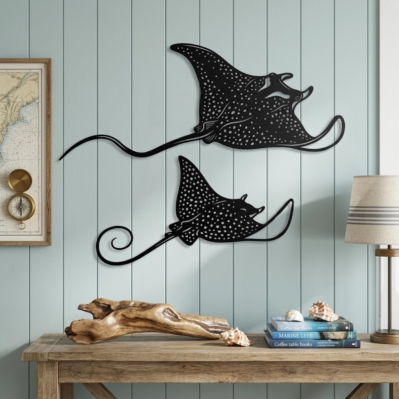May include: Two black metal wall art pieces shaped like spotted eagle rays. Each ray features a flat silhouette with intricate cut-out details representing spots across the body and wings, and a long, thin, curved tail extending from the main body.