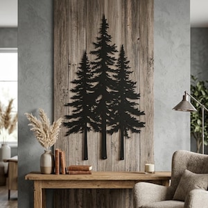 May include: A black metal wall art piece featuring a silhouette of three pine trees mounted on a vertical wooden plank background. The design displays detailed needle textures and branches, creating a sharp contrast against the light brown wood grain.