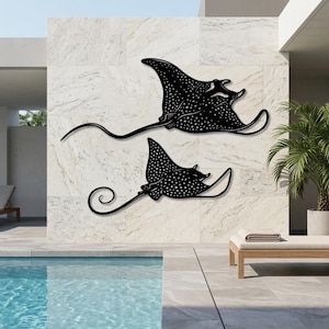May include: Two black metal wall art pieces shaped like spotted eagle rays. Each ray features a flat silhouette with a long, slender tail and a distinct pattern of white dots across the body, mounted horizontally on a light-colored stone wall.