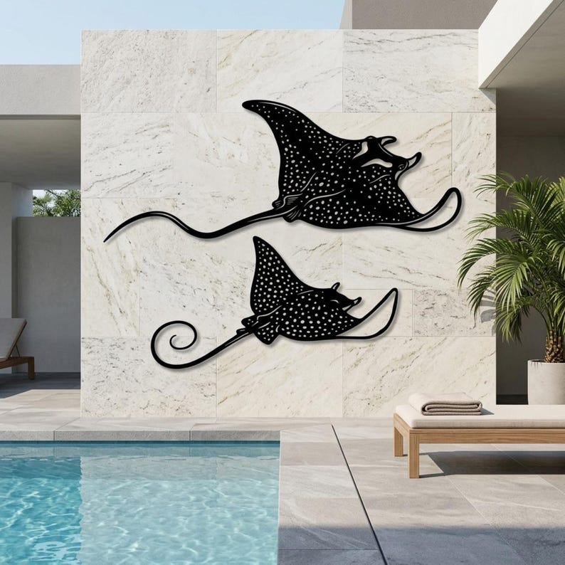 May include: Two black metal wall art pieces shaped like spotted eagle rays. Each ray features a flat silhouette with a long, slender tail and a distinct pattern of white dots across the body, mounted horizontally on a light-colored stone wall.