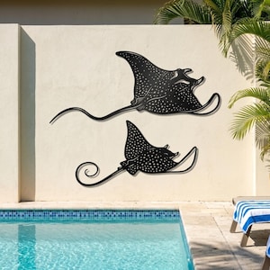 May include: Two black metal wall art pieces shaped like spotted eagle rays. Each ray features a flat silhouette with intricate white dot patterns across the body, extended wing-like fins, and a long, thin, curved tail attached to the wall.