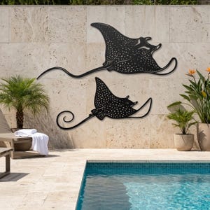 May include: Two black metal wall art pieces shaped like spotted eagle rays. Each silhouette features a flat body with a long, thin, curved tail and a detailed pattern of white dots across the surface, mounted horizontally on a stone wall.