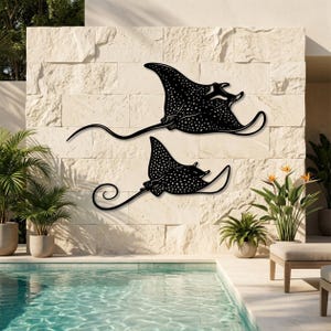 May include: Two black metal wall art pieces shaped like spotted eagle rays, featuring intricate white dot patterns across their bodies and long, slender, curved tails. These flat, silhouette-style marine animal decorations are mounted on a stone wall.