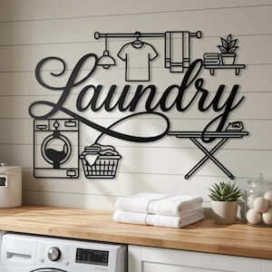 Pode incluir: Uma placa de parede em metal preto com a palavra Laundry em letra cursiva. O design inclui ícones de uma máquina de lavar, um cesto de roupa suja com peças, uma camisa pendurada, uma toalha, uma prateleira com uma planta e uma tábua de passar.