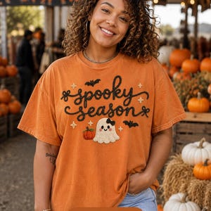 Puede incluir: Una camiseta naranja de manga corta con el texto bordado en negro spooky season, una pequeña calabaza naranja, un fantasma blanco con un lazo negro, y murciélagos y estrellas negras. La prenda tiene un cuello redondo.