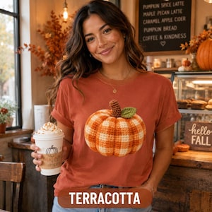 Puede incluir: Una camiseta de color terracota con un gráfico grande de una calabaza de ganchillo a cuadros naranjas y blancos, con un tallo marrón y una hoja verde. La camiseta se lleva con un vaso de café blanco que tiene una ilustración de calabaza.