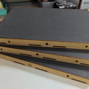 Könnte beinhalten: Drei rechteckige Tabletts, die übereinander gestapelt sind, jedes mit einem hellbraunen Holzrahmen mit sichtbaren Fingerzinken und einer strukturierten dunkelgrauen Schaumstoffeinlage auf der Oberseite sowie kleinen Löchern und Schlitzen.