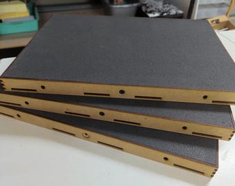 Op de afbeelding: Drie rechthoekige dienbladen die op elkaar zijn gestapeld, elk met een lichtbruin houten frame met zichtbare vingerverbindingen en een gestructureerde donkergrijze schuiminzet op de bovenkant, met kleine ronde gaten en sleuven aan de zijkanten.