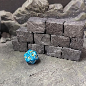 May include: A stack of ten dark grey, rectangular stone-like blocks arranged in a wall formation, positioned next to a single, twenty-sided, bright blue polyhedral die with gold-coloured numbers engraved on its triangular faces.