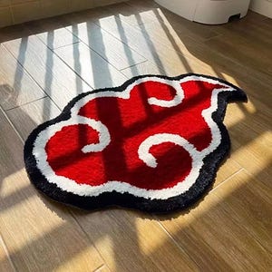 May include: A red cloud-shaped rug with white and black accents. The rug features a bold design with a red center, white swirling details, and a black border. The rug is placed on a wooden floor.