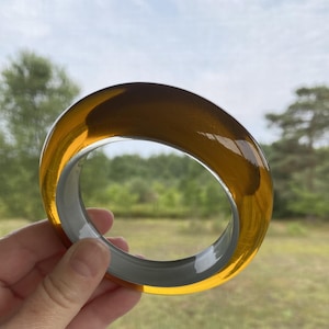 May include: A thick, circular bangle bracelet featuring a two-tone design. The outer portion is a translucent, warm amber-colored material, while the inner portion is a solid, opaque gray material, creating a distinct contrast in color and opacity.