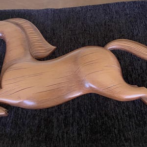 Puede incluir: Una escultura de pared de un caballo hecha de madera de color marrón claro con un acabado suave y pulido. El caballo se muestra en una pose dinámica al galope con la cabeza girada, crines fluidas y patas extendidas, mostrando la veta.
